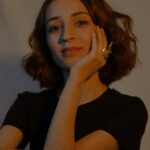 A sophisticated portrait of a woman with wavy hair wearing a t-shirt and ring indoors.