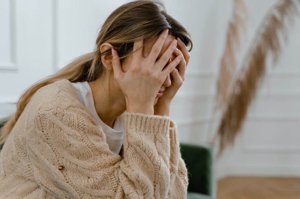 holistic wellness therapy for past trauma A woman sitting indoors covering her face in frustration, depicting stress and mental health challenges.